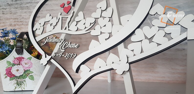 May include: White wooden heart-shaped sign with a clear acrylic insert. The sign features a couple's names, "Jeri and Chase", and their wedding date, "5-4-2019". The sign is decorated with small white hearts and a black border.