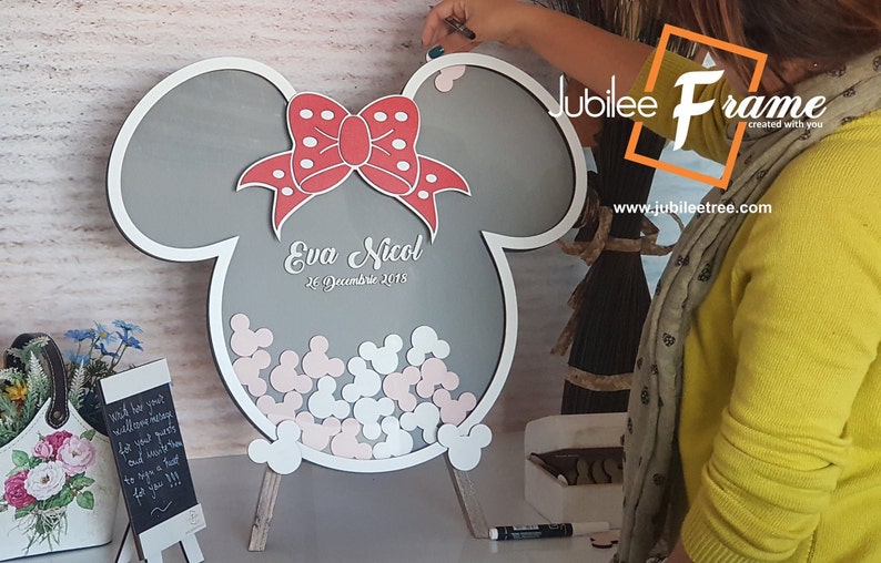 May include: A gray wooden Mickey Mouse shaped sign with a red bow and white polka dots. The sign says "Eva Nicol 26 December 2015" and has a space for guests to leave their well wishes.