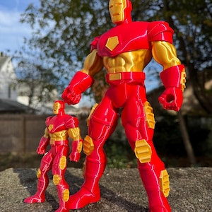 Puede incluir: Dos figuras de acci&oacute;n de Iron Man rojas y doradas. La figura m&aacute;s grande est&aacute; de pie con los brazos a los lados. La figura m&aacute;s peque&ntilde;a est&aacute; de pie con los brazos ligeramente doblados en los codos. Ambas figuras tienen un brillo met&aacute;lico.