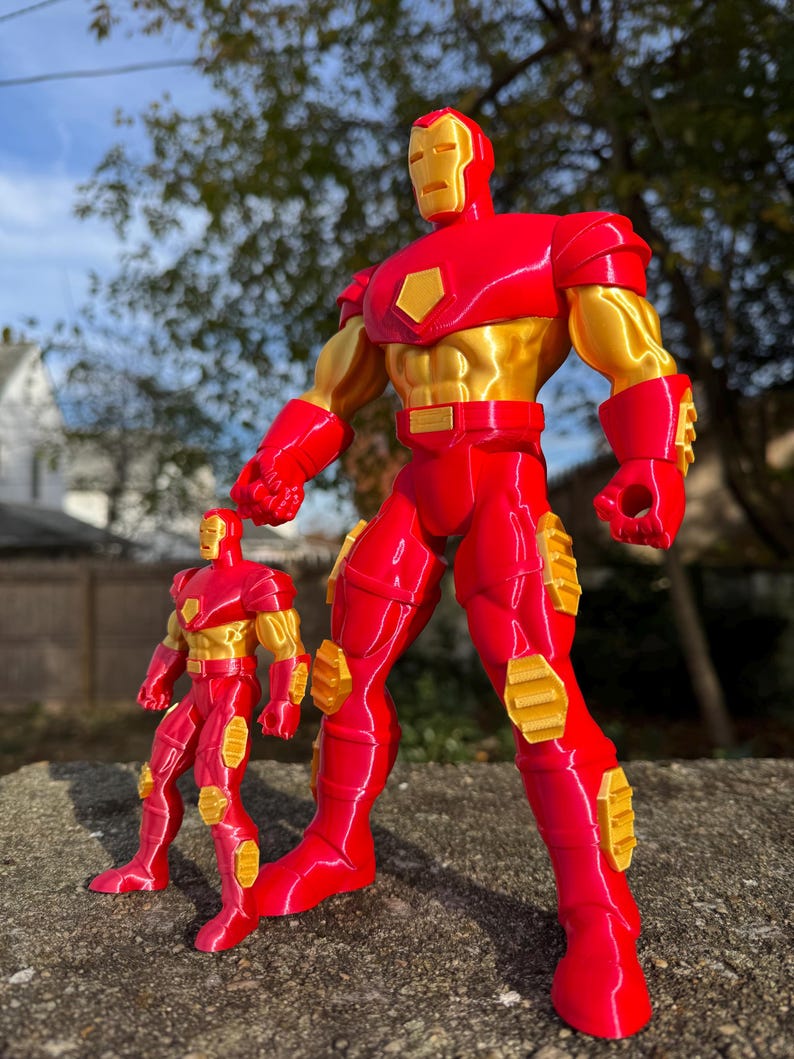 Puede incluir: Dos figuras de acci&oacute;n de Iron Man rojas y doradas. La figura m&aacute;s grande est&aacute; de pie con los brazos a los lados. La figura m&aacute;s peque&ntilde;a est&aacute; de pie con los brazos ligeramente doblados en los codos. Ambas figuras tienen un brillo met&aacute;lico.