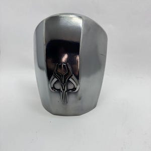 Op de afbeelding: Een zilverkleurige, gebogen helm met een gepolijste afwerking. De helm heeft een gedetailleerd embleem op de voorkant, met een gestileerd, symmetrisch ontwerp. De algehele esthetiek suggereert een futuristisch of sciencefictionthema.