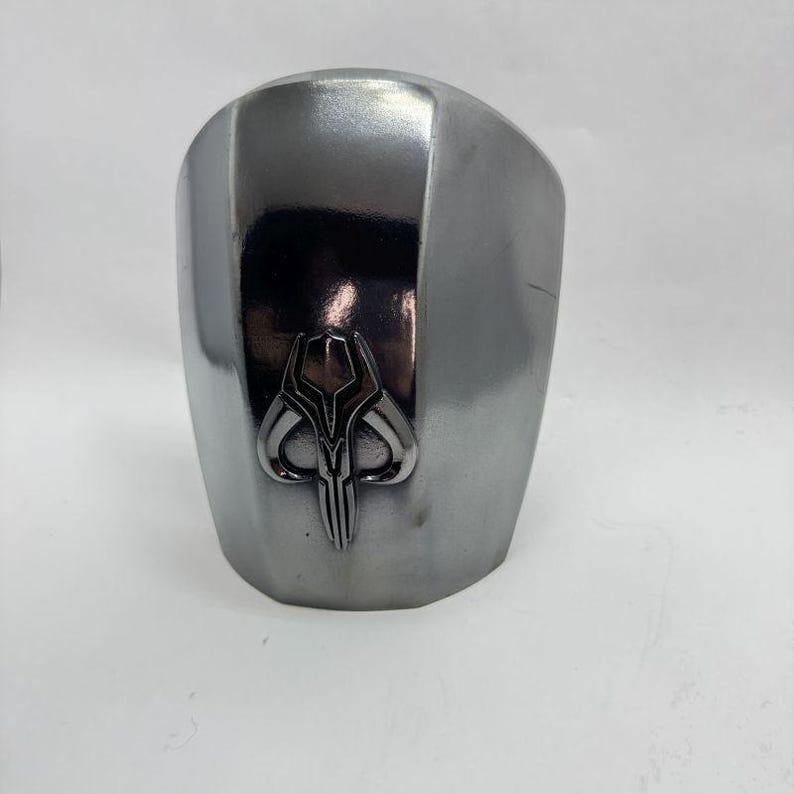 Op de afbeelding: Een zilverkleurige, gebogen helm met een gepolijste afwerking. De helm heeft een gedetailleerd embleem op de voorkant, met een gestileerd, symmetrisch ontwerp. De algehele esthetiek suggereert een futuristisch of sciencefictionthema.