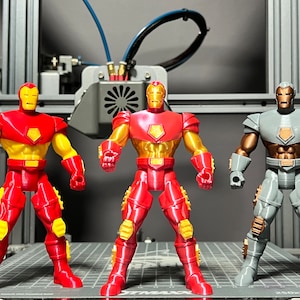 Puede incluir: Tres figuras de acci&oacute;n, dos figuras de Iron Man rojas y doradas y una figura de Iron Man gris y dorada. Las figuras est&aacute;n de pie sobre una superficie de rejilla gris y negra.