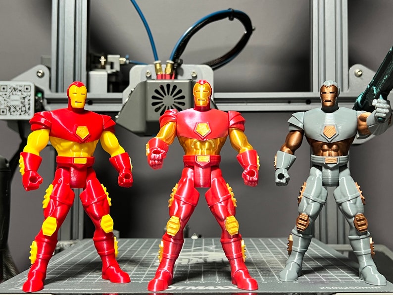Puede incluir: Tres figuras de acci&oacute;n, dos figuras de Iron Man rojas y doradas y una figura de Iron Man gris y dorada. Las figuras est&aacute;n de pie sobre una superficie de rejilla gris y negra.