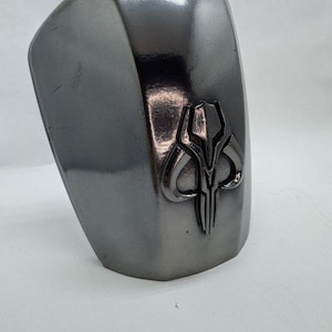 Op de afbeelding: Een metalen grijze schouderpantser met een verhoogd embleem. Het embleem is een gestileerde schedel met hoorns en verticale lijnen. Het pantser heeft een gebogen vorm en een glad, reflecterend oppervlak.