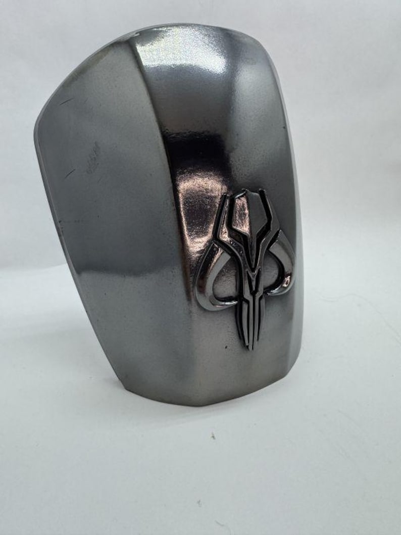 Op de afbeelding: Een metalen grijze schouderpantser met een verhoogd embleem. Het embleem is een gestileerde schedel met hoorns en verticale lijnen. Het pantser heeft een gebogen vorm en een glad, reflecterend oppervlak.