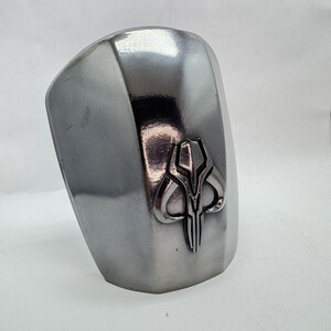 Op de afbeelding: Een zilverkleurig, gebogen pantserstuk met een verhoogd, gestileerd embleem. Het embleem is een gedetailleerd, symmetrisch ontwerp in een donkerdere zilverkleur, waarschijnlijk een symbool van trouw of affiliatie.