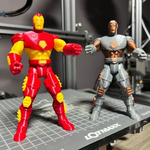 Puede incluir: Dos figuras de acci&oacute;n de Iron Man impresas en 3D. Una es roja y amarilla, la otra es gris y marr&oacute;n. Ambas figuras est&aacute;n de pie sobre una superficie de rejilla gris.