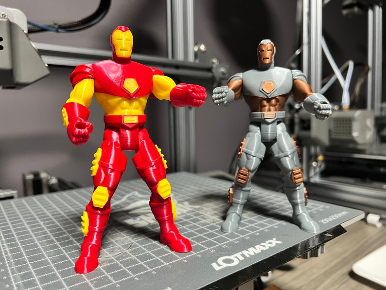 Puede incluir: Dos figuras de acci&oacute;n de Iron Man impresas en 3D. Una es roja y amarilla, la otra es gris y marr&oacute;n. Ambas figuras est&aacute;n de pie sobre una superficie de rejilla gris.