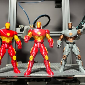 Puede incluir: Tres figuras de acci&oacute;n, dos figuras de Iron Man rojas y doradas y una figura de Iron Man gris y marr&oacute;n con una pistola azul. Las figuras est&aacute;n de pie sobre una plataforma gris.
