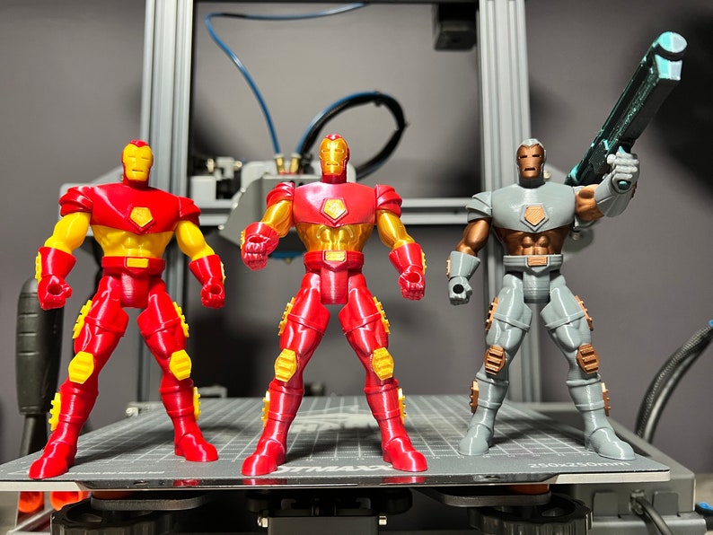 Puede incluir: Tres figuras de acci&oacute;n, dos figuras de Iron Man rojas y doradas y una figura de Iron Man gris y marr&oacute;n con una pistola azul. Las figuras est&aacute;n de pie sobre una plataforma gris.