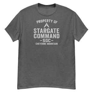 May include: A dark gray t-shirt with the text "PROPERTY OF STARGATE COMMAND - SGC - CHEYENNE MOUNTAIN" printed in white. The shirt has a crew neck and short sleeves. The text is in a distressed, vintage style.