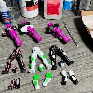 Puede incluir: Piezas de figuras de acci&oacute;n de pl&aacute;stico en colores rojo, blanco y negro. Las piezas incluyen piernas, brazos y manos. Las piezas est&aacute;n dispuestas sobre una superficie de madera.