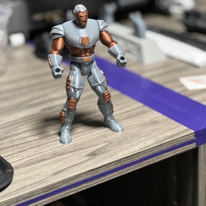 Puede incluir: Una figura de acci&oacute;n de superh&eacute;roe gris y cobre con un casco y una placa de pecho. La figura est&aacute; de pie sobre una superficie de madera.