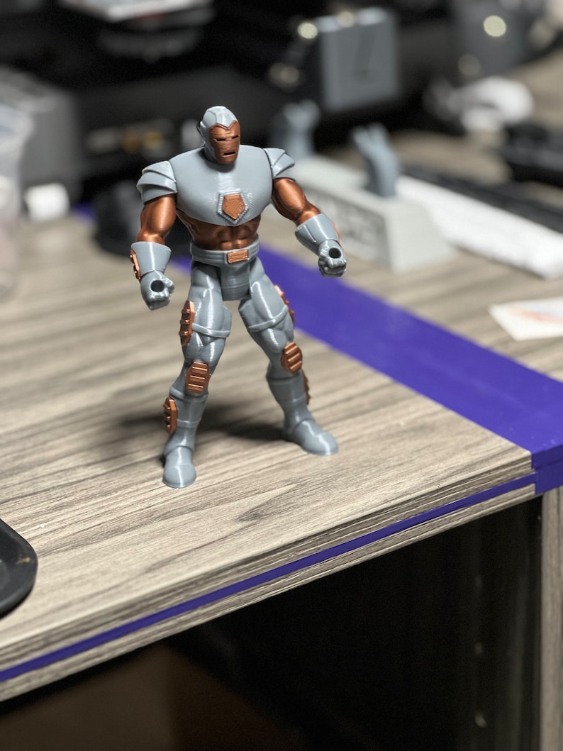 Puede incluir: Una figura de acci&oacute;n de superh&eacute;roe gris y cobre con un casco y una placa de pecho. La figura est&aacute; de pie sobre una superficie de madera.