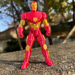 Puede incluir: Una figura de acci&oacute;n de Iron Man roja y dorada con un dise&ntilde;o detallado. La figura est&aacute; de pie sobre una superficie texturizada.
