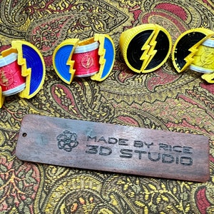 May include: A collection of 3D-printed Flash-themed accessories in yellow, blue, red, and black. The items include rings and a wooden tag that reads "MADE BY RICE 3D STUDIO". The accessories are displayed on a patterned fabric.