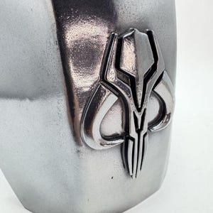 Op de afbeelding: Close-up van een zilverkleurig metalen object met een gedetailleerd embleem. Het embleem heeft een symmetrisch ontwerp met een centraal verticaal element en gebogen vormen. Het object heeft een getextureerd oppervlak.