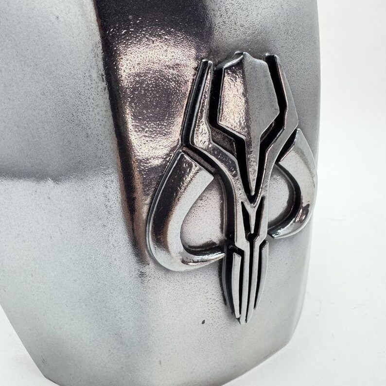 Op de afbeelding: Close-up van een zilverkleurig metalen object met een gedetailleerd embleem. Het embleem heeft een symmetrisch ontwerp met een centraal verticaal element en gebogen vormen. Het object heeft een getextureerd oppervlak.