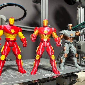 Puede incluir: Tres figuras de acci&oacute;n, dos figuras de Iron Man rojas y amarillas y una figura gris con una pistola verde. Las figuras est&aacute;n de pie sobre una superficie gris con un patr&oacute;n de cuadr&iacute;cula.