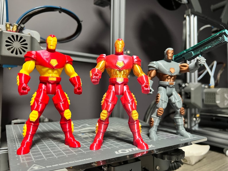 Puede incluir: Tres figuras de acci&oacute;n, dos figuras de Iron Man rojas y amarillas y una figura gris con una pistola verde. Las figuras est&aacute;n de pie sobre una superficie gris con un patr&oacute;n de cuadr&iacute;cula.