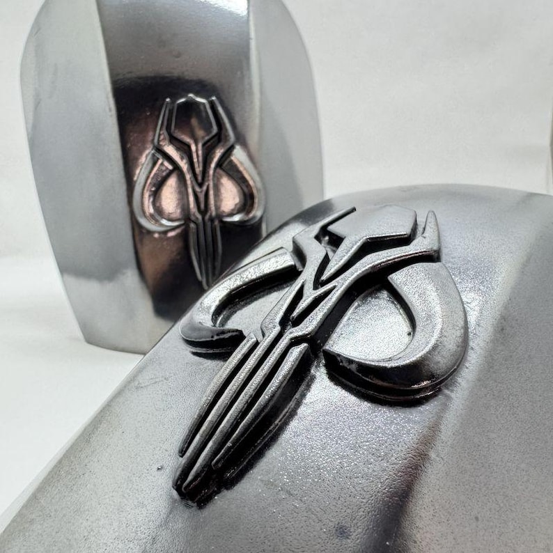 Op de afbeelding: Close-up van twee zilverkleurige Mandalorian pantserstukken. Elk stuk heeft een verhoogd, gedetailleerd embleem in het midden. Het embleem is een gestileerd ontwerp met een centrale vorm en gebogen elementen.