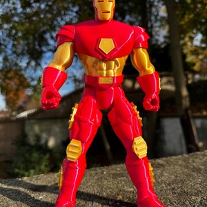 Puede incluir: Una figura de acci&oacute;n de Iron Man impresa en 3D de color rojo y dorado. La figura est&aacute; de pie con los brazos a los lados y los pu&ntilde;os cerrados. La figura tiene un dise&ntilde;o detallado con un acabado met&aacute;lico.