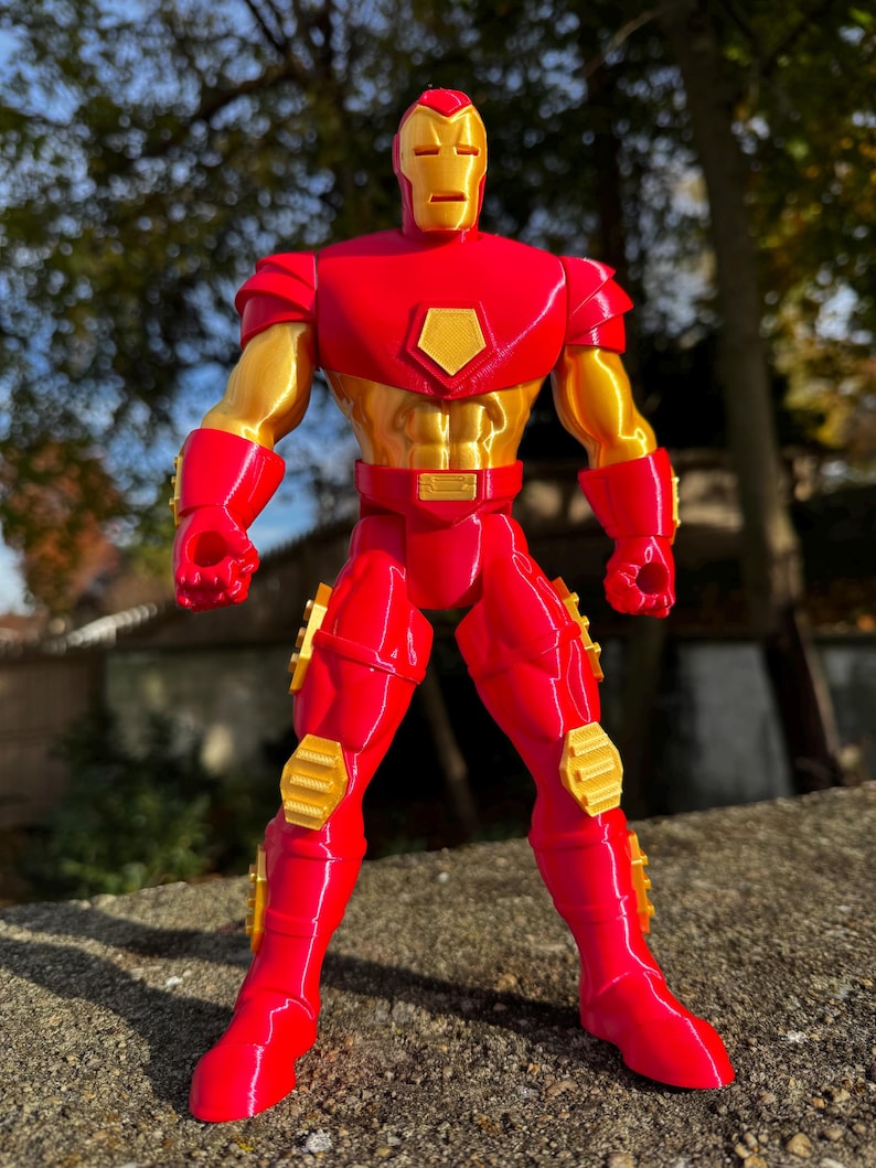 Puede incluir: Una figura de acci&oacute;n de Iron Man impresa en 3D de color rojo y dorado. La figura est&aacute; de pie con los brazos a los lados y los pu&ntilde;os cerrados. La figura tiene un dise&ntilde;o detallado con un acabado met&aacute;lico.
