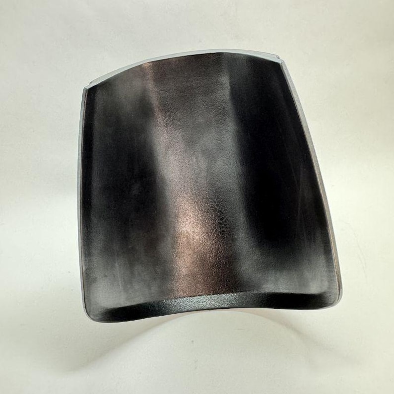 Op de afbeelding: Een donkergrijs, gebogen metalen object met een rechthoekige vorm en afgeronde hoeken. De binnenkant heeft een textuur en de randen zijn omlijnd met een lichter grijs. Het object is waarschijnlijk een onderdeel van een groter item.