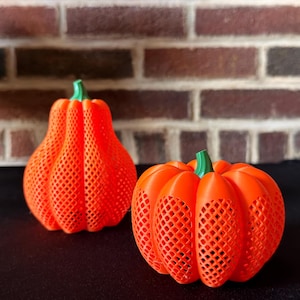 May include: Two orange 3D-printed pumpkins with green stems. The pumpkins have a lattice design and sit on a black surface in front of a brick wall. One pumpkin is pear-shaped, and the other is round.