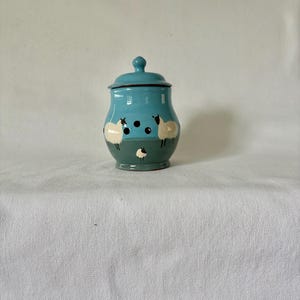 May include: A teal and green ceramic jar with a lid, decorated with sheep motifs. The sheep are white with black details. The jar has a glossy finish. Measurements are in centimetres.