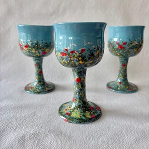 May include: Three ceramic goblets featuring a light blue upper bowl and a stem decorated with a hand-painted meadow scene. The design includes small red poppies, yellow wildflowers, and green foliage that extends from the base up to the bowl.