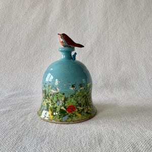 May include: A turquoise ceramic bell with a painted landscape design and a small robin on top. The landscape features green, yellow, and red accents. The robin is brown and orange.