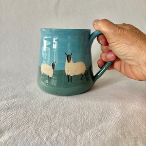 May include: A ceramic mug with a glossy finish featuring a two-tone design of light blue on the top half and dark teal on the bottom half. The mug is decorated with hand-painted illustrations of two cream-coloured sheep with black faces and legs.