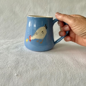 May include: A blue ceramic mug featuring a white chicken design. The mug has a curved handle and a light blue interior. The chicken has black, yellow, and red details. The mug is held by a hand.