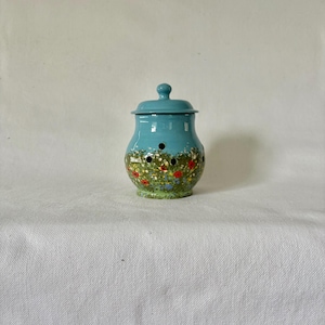 May include: A turquoise ceramic garlic keeper with a lid. The jar has a floral design with red, orange, and green accents. It has three small holes for ventilation. The lid has a small knob on top.