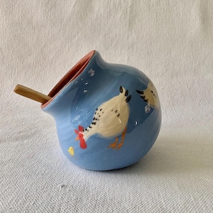 May include: A blue ceramic salt cellar with a chicken illustration and a small wooden spoon. The interior is coral. The chicken is white with black spots and a red comb. A yellow chick is also painted on the side.