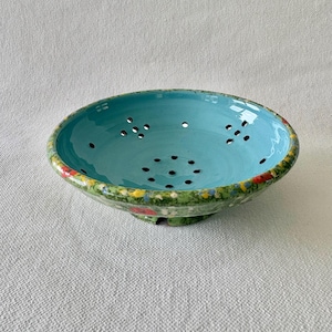 May include: A shallow ceramic colander featuring a vibrant turquoise interior and a green exterior rim decorated with small, colourful floral patterns. The bowl has multiple drainage holes distributed across the base and sides for straining.