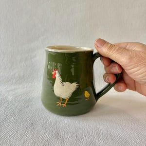 May include: A green ceramic mug featuring a white chicken and a yellow chick illustration. The mug has a rounded shape, a handle, and a cream-coloured interior. The chicken has red accents. The chick is small and yellow.