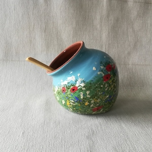 May include: A blue ceramic jar with a floral design featuring red poppies and green foliage. The jar has a wide opening and a wooden spoon resting on the rim.