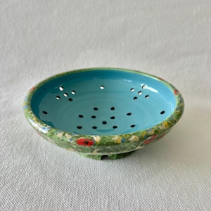 May include: A small ceramic colander with a bright turquoise interior and a green exterior rim. The rim is decorated with colourful floral patterns, including red poppies. The colander has small drainage holes and is set against a white background.