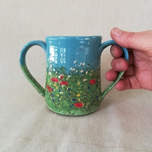 May include: A ceramic mug with two handles, decorated with a meadow design. The top is bright blue, fading to green with red poppies, white and yellow flowers. The mug is held by a hand.