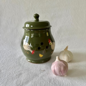 May include: A green ceramic garlic keeper with a lid, decorated with painted chickens and a chick. Two garlic bulbs are next to the keeper. The keeper has ventilation holes.