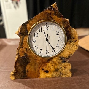 May include: A wooden clock with a natural wood grain finish. The clock face is white with black numbers and hands. The clock is shaped like a house and is sitting on a brown surface.