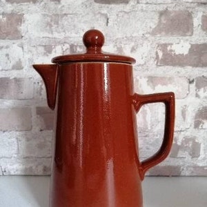 May include: A glazed, reddish-brown ceramic coffee pot with a lid and handle. The pot features a pouring spout and a rounded knob on the lid. The surface is shiny, reflecting light. The background is a brick wall.