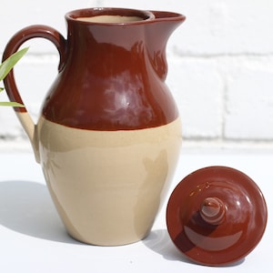 May include: A brown and beige ceramic jug with a lid. The jug has a wide mouth and a handle. The lid is round and has a knob on top.