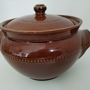 May include: A brown ceramic casserole dish with a lid. The dish has two handles and a decorative beaded pattern around the base. The lid has a knob and a decorative pattern. The dish is glazed and has a glossy finish.