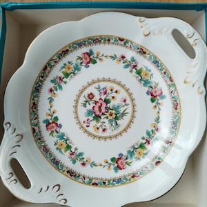 May include: White porcelain serving plate with a scalloped edge and gold trim. The centre features a floral design with pink, yellow, and blue flowers. The plate has two handles and is in a blue box.