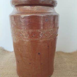 May include: A brown stoneware jar with a cylindrical shape and a slightly flared rim. The jar features a decorative band of embossed floral and linear patterns around the middle. The surface has a speckled appearance.