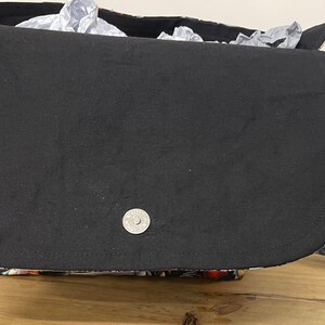 Puede incluir: Un bolso bandolera de tela negra con cierre de solapa y un bot&oacute;n a presi&oacute;n plateado. El bolso tiene un estampado floral en la parte posterior y una correa negra.